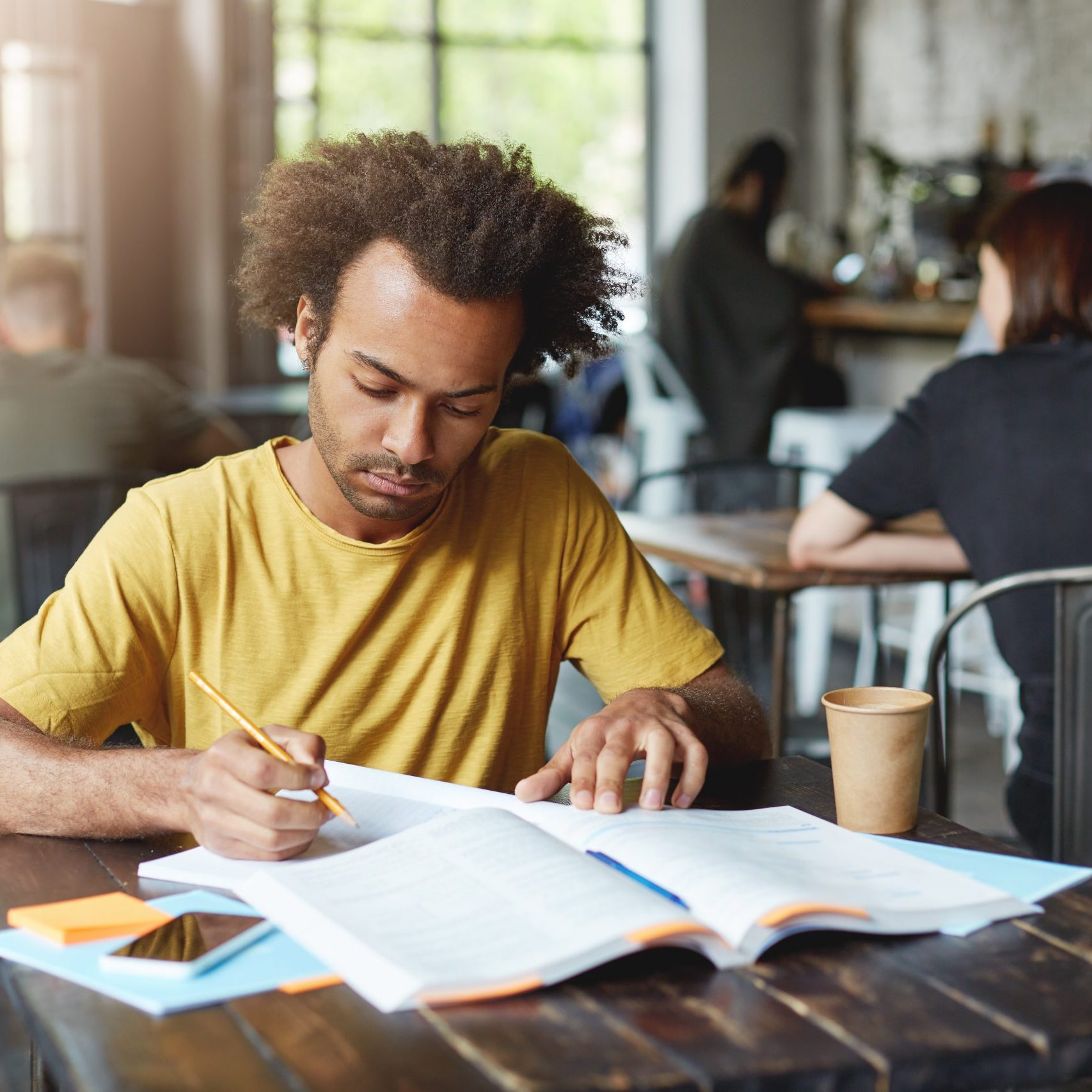 Close-up portrait of serious dark-skinned male student wearing yellow T-shirt sitting at cafe during break drinking coffee and preparing for lessons writing in copybook from book with pencil