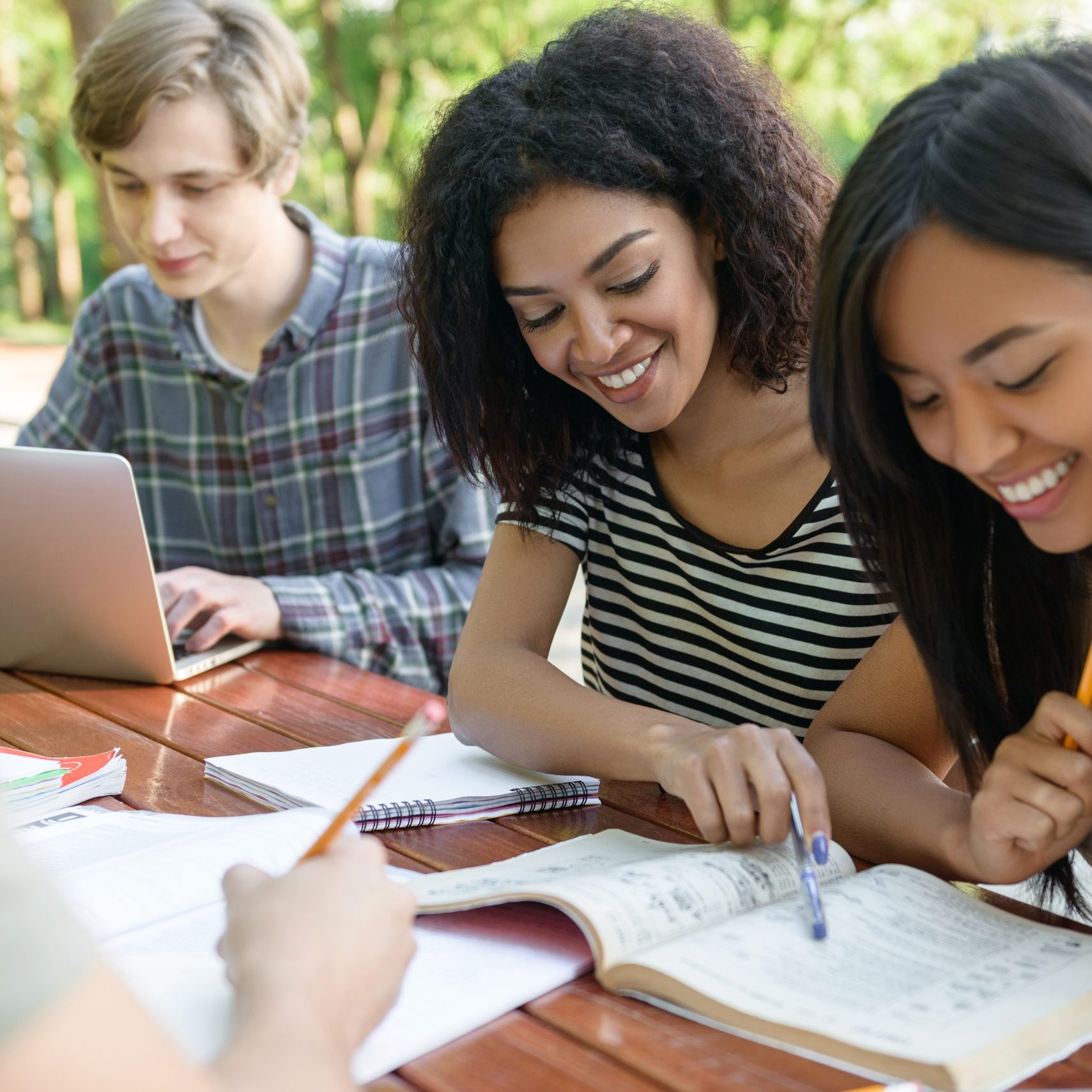 Image of multiethnic group of young happy students sitting and studying outdoors while talking. Looking aside.